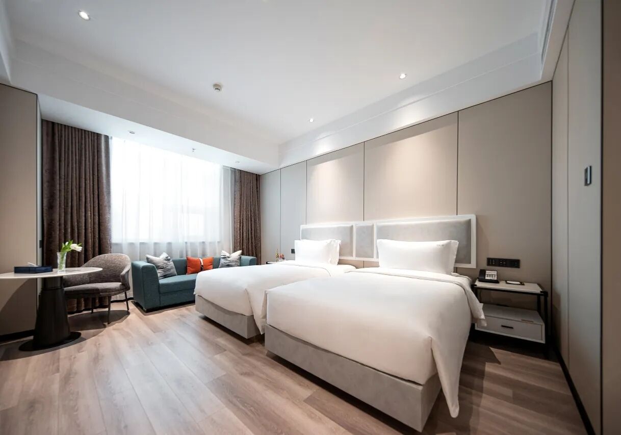 A modern hotel room with two beds, a sofa, a desk, and a large window with curtains.