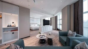 Una habitación de hotel moderna con una cama grande, una zona de estar con sofá y mesa de centro, un televisor y un ventanal con vistas a la ciudad.