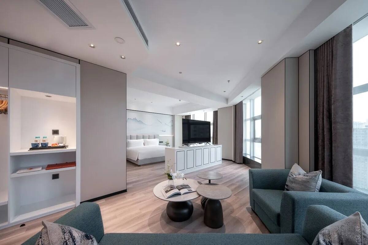 Une chambre d’hôtel moderne dotée d’un grand lit, d’un coin salon avec un canapé et une table basse, d’une télévision et d’une grande fenêtre donnant sur la ville.
