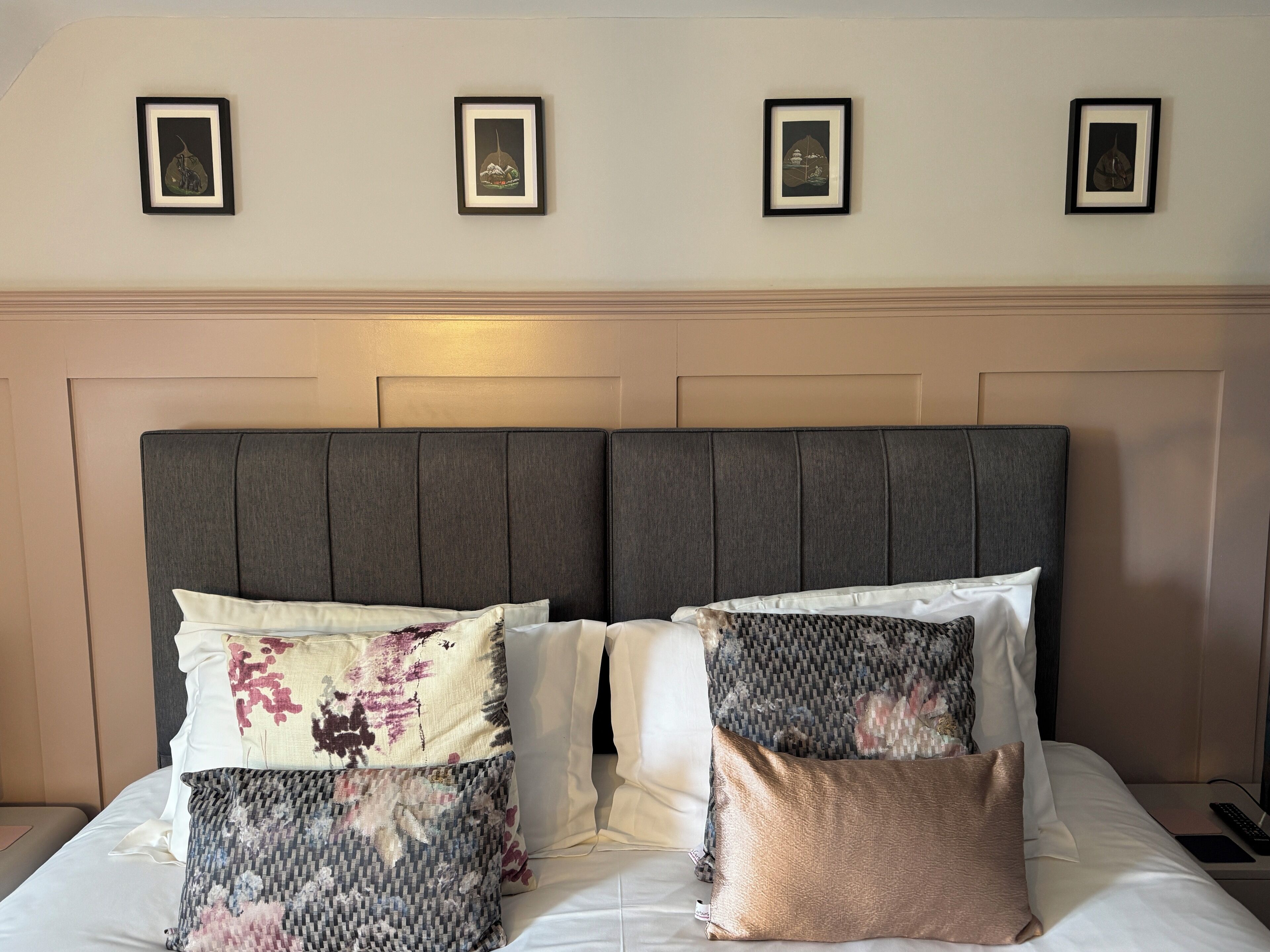 Una cama con cabecera tapizada en gris, varios almohadones y tres cuadros en la pared superior.
