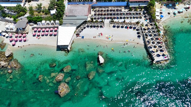 An aerial view of a beach with clear turquoise water, rocky outcrops, and a pavilion with umbrellas. - La Terrazza Azzurra (Vlore)