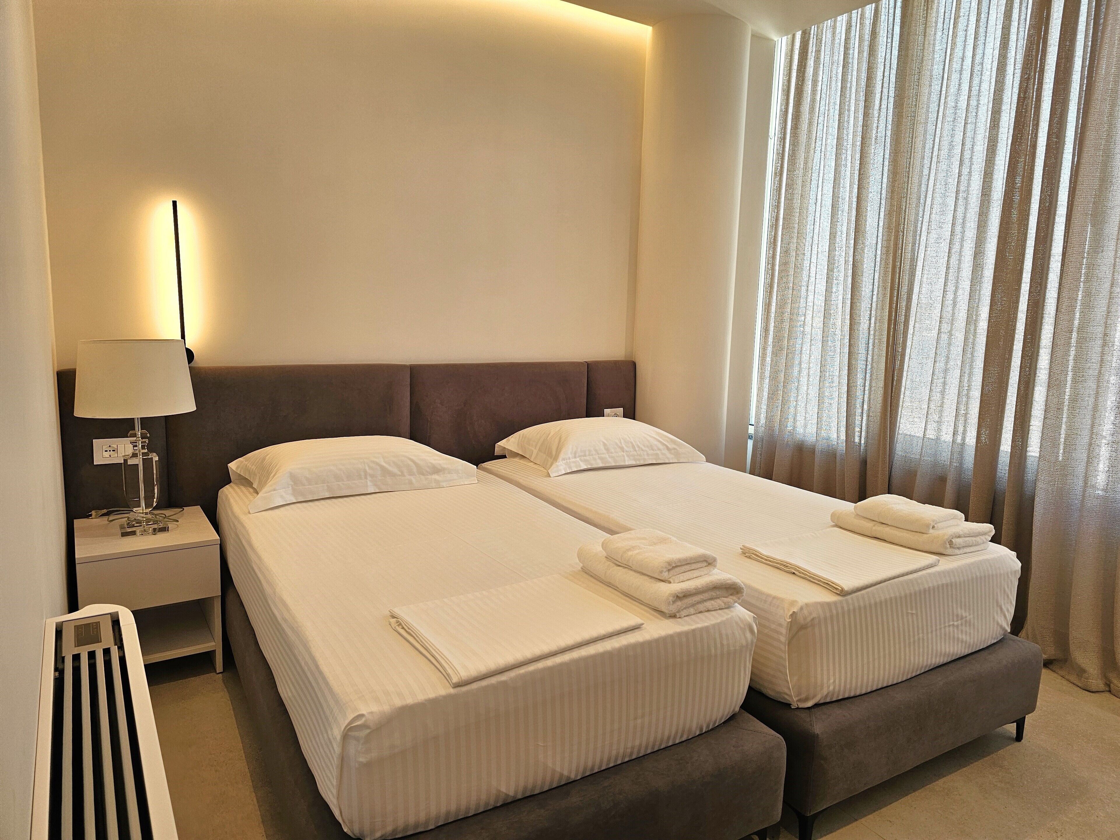 A hotel room with two beds, a nightstand with a lamp, and a window with curtains.