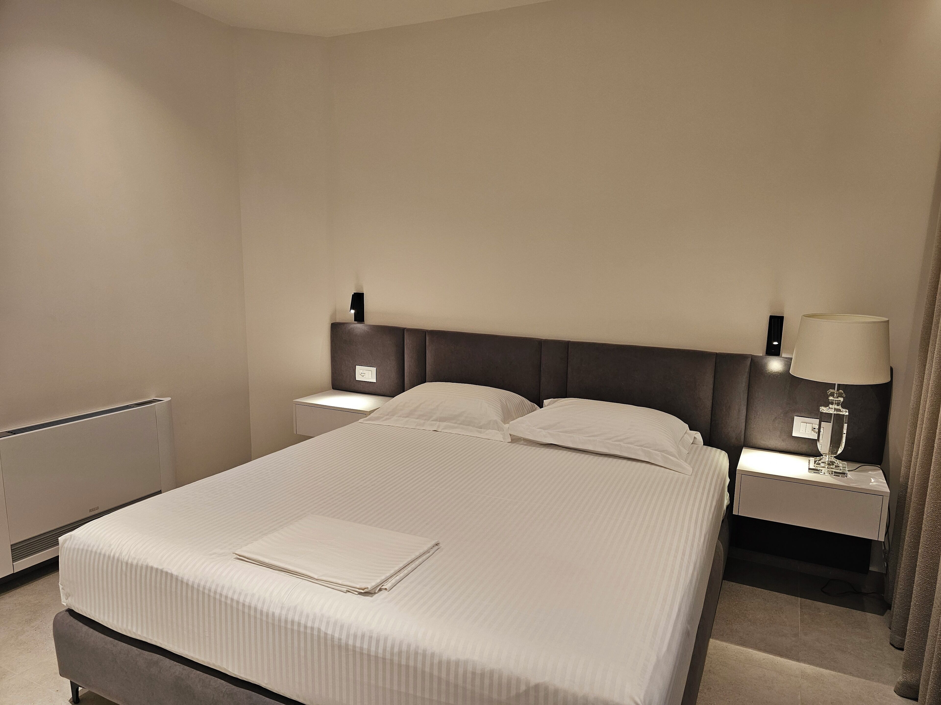 Ein ordentlich gemachtes Bett mit einem gefalteten Handtuch, ein Nachttisch mit einer Lampe und eine Wandleuchte.
