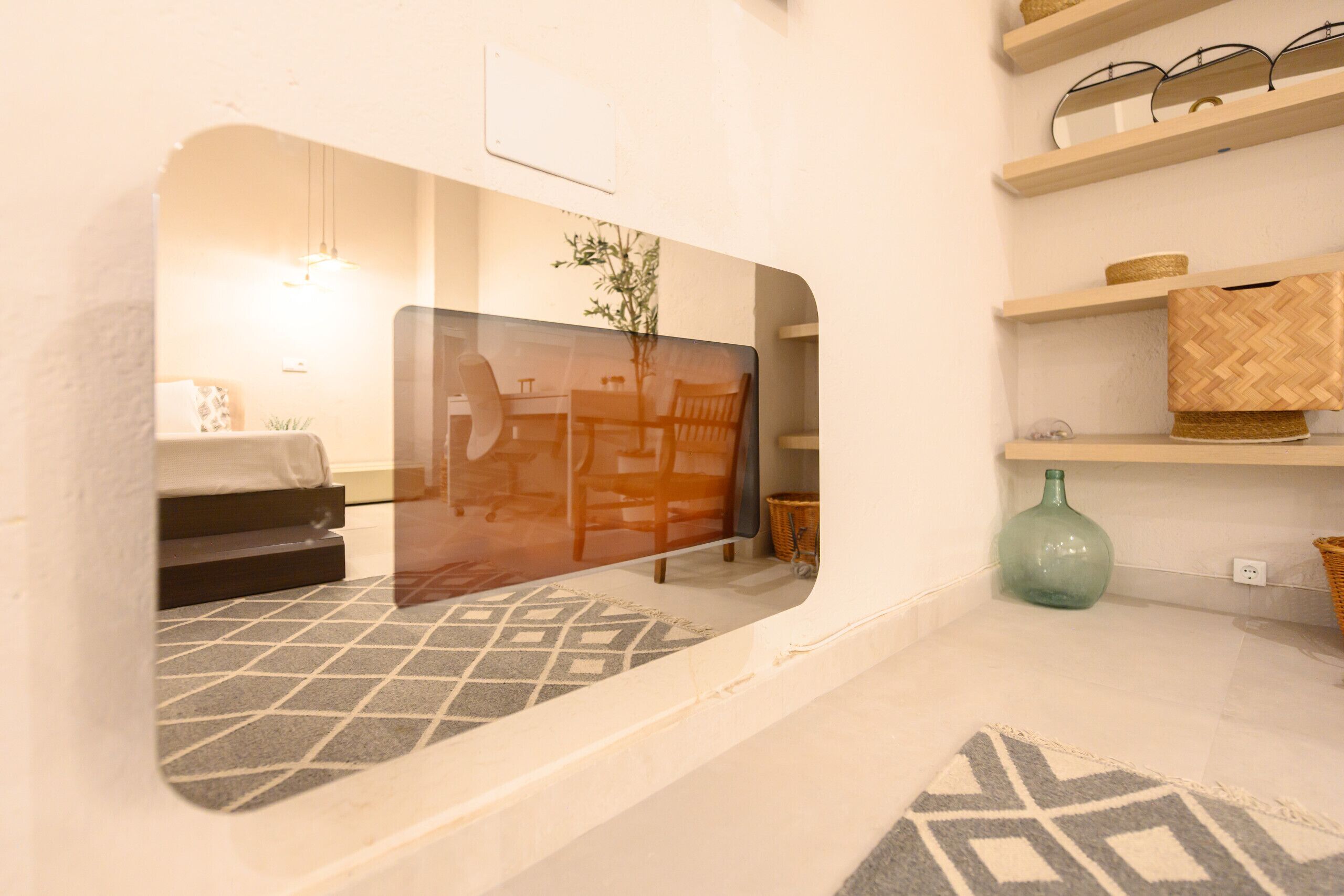 Una habitación con pared espejada, alfombra estampada, un jarrón verde y estantes con cestas.