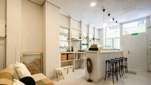 Une pièce accueillante avec un canapé, un petit tabouret, un coin bar avec des tabourets, des étagères remplies d’objets et une porte portant l’inscription « SORTIE ».