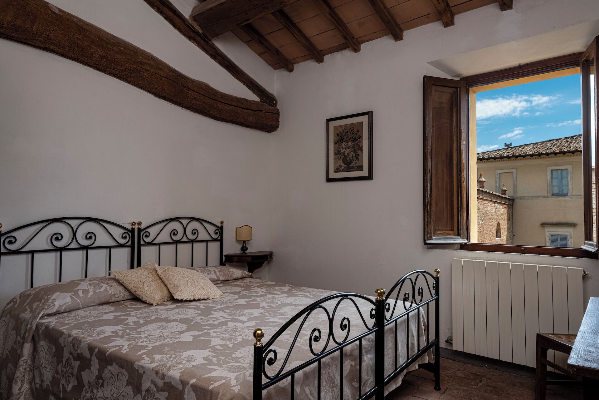 Een slaapkamer met een bed, een nachtkastje, een raam met uitzicht op gebouwen en een lijst met een afbeelding aan de muur.
