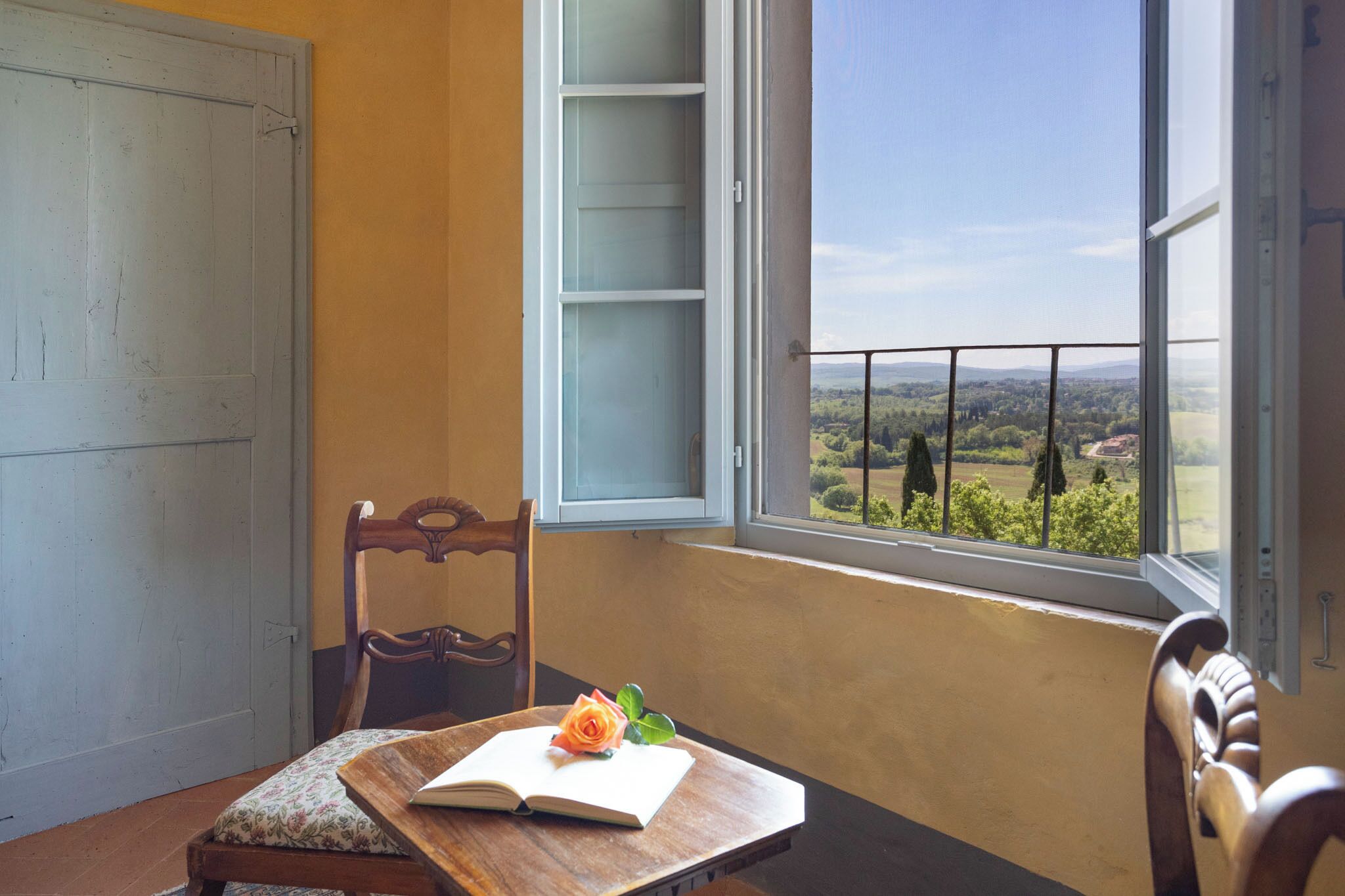 A room with a wooden table, a chair, an open book, and a window with a view of greenery.