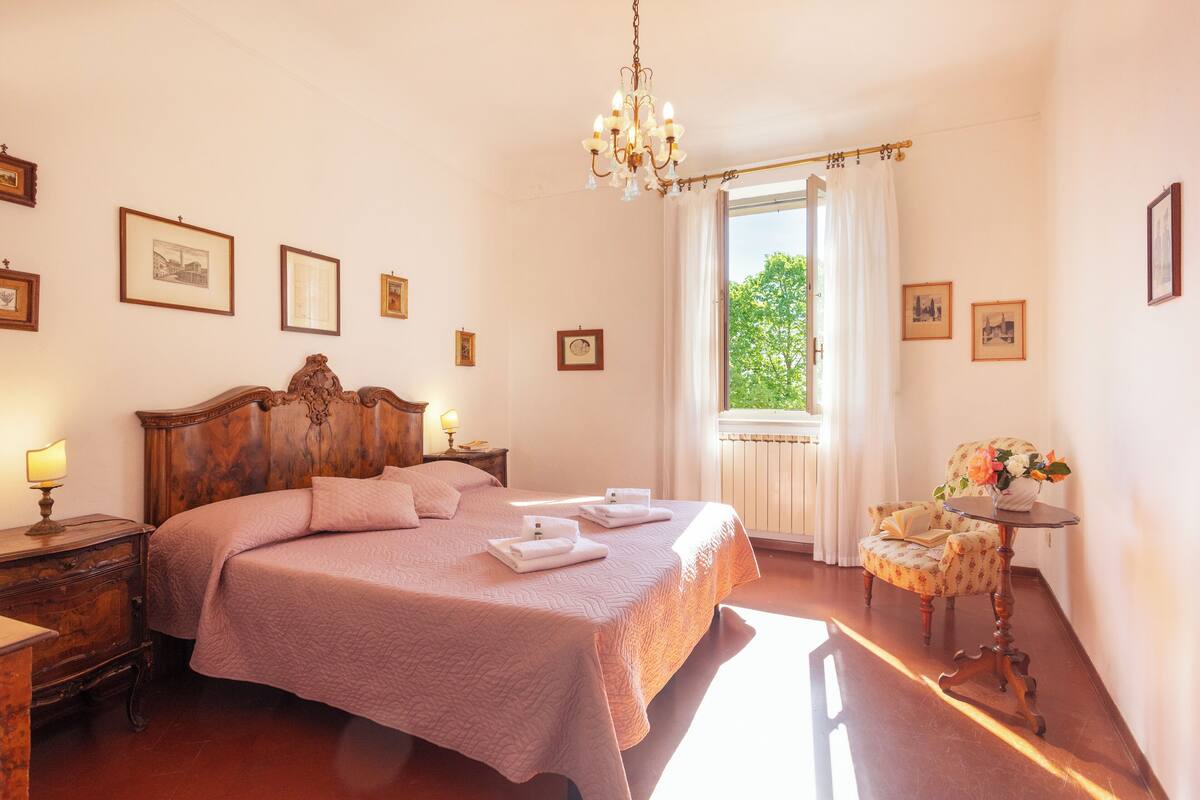 A bedroom with a large bed, a wooden nightstand, a chair, and a table with a vase of flowers.