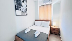 A neatly made bed with white linens and two rolled towels, a wooden nightstand with a lamp, a wall-mounted air conditioner, and a large abstract painting. - Downtown Bliss – Piraeus Street by ΜΡS (Athina)