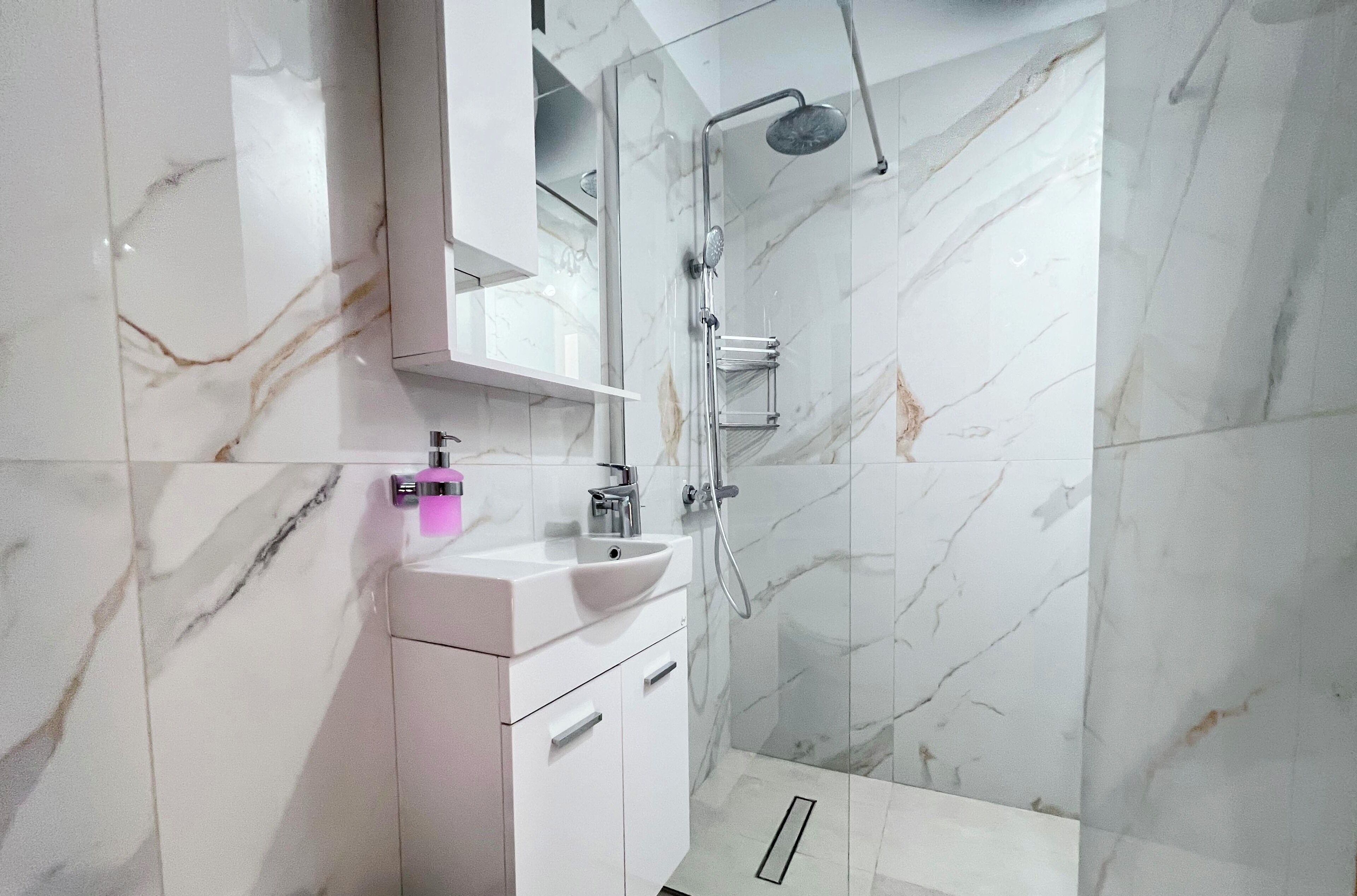 A modern bathroom with a marble wall, a white sink, and a glass-enclosed shower.