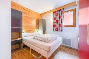 A bedroom with a bed, wooden panel wall, window with red curtains, and built-in shelving.
