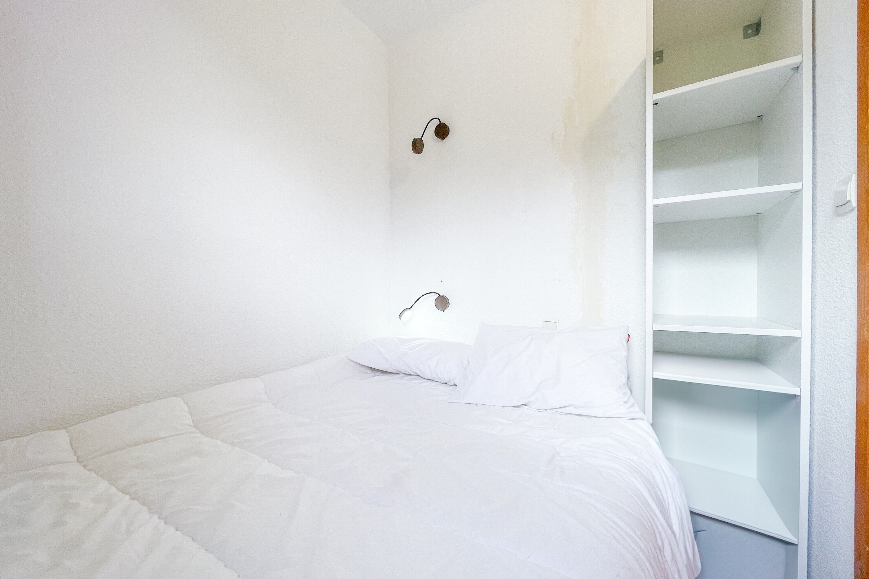 A white bed with a headboard, a wall-mounted light fixture, and a white shelving unit.