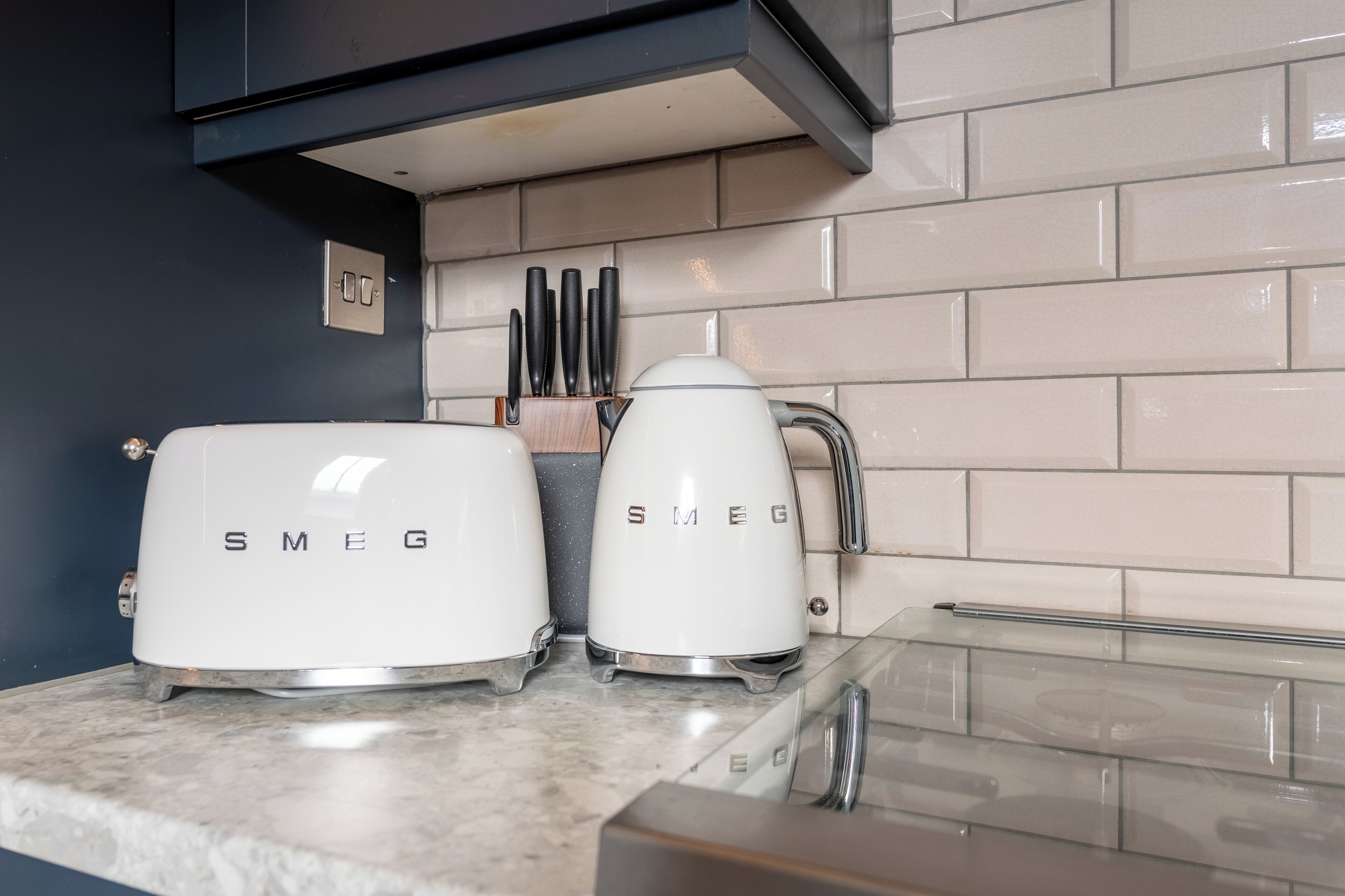 Two white Smeg electric kettles on a kitchen counter with a tiled backsplash and a black wall.