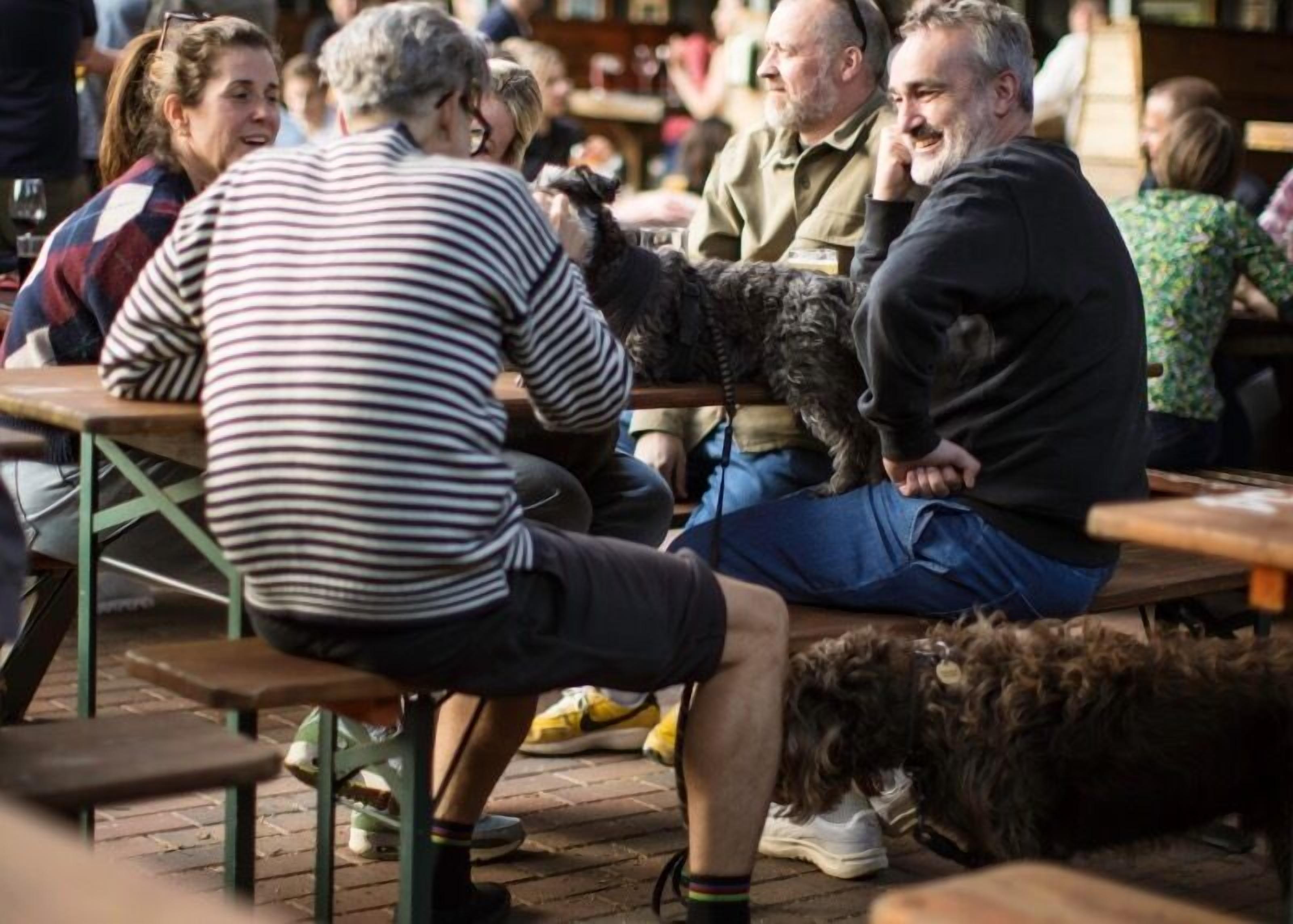 Personas sentadas en mesas de madera al aire libre, algunas acompañadas por perros.