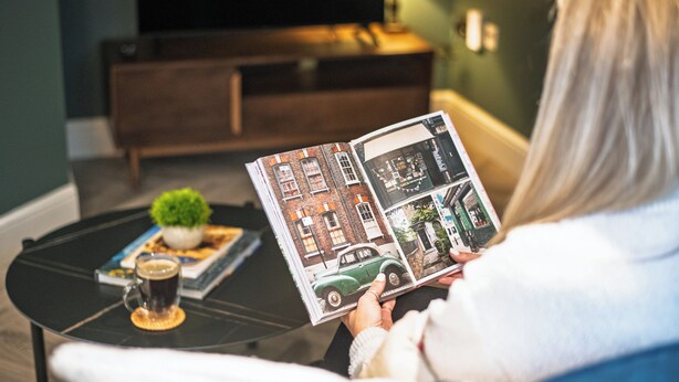 A person reading a magazine on a black coffee table with a small plant and a cup of coffee.