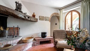 A cozy room with a fireplace, a comfortable chair, a table, and a view through an arched window.