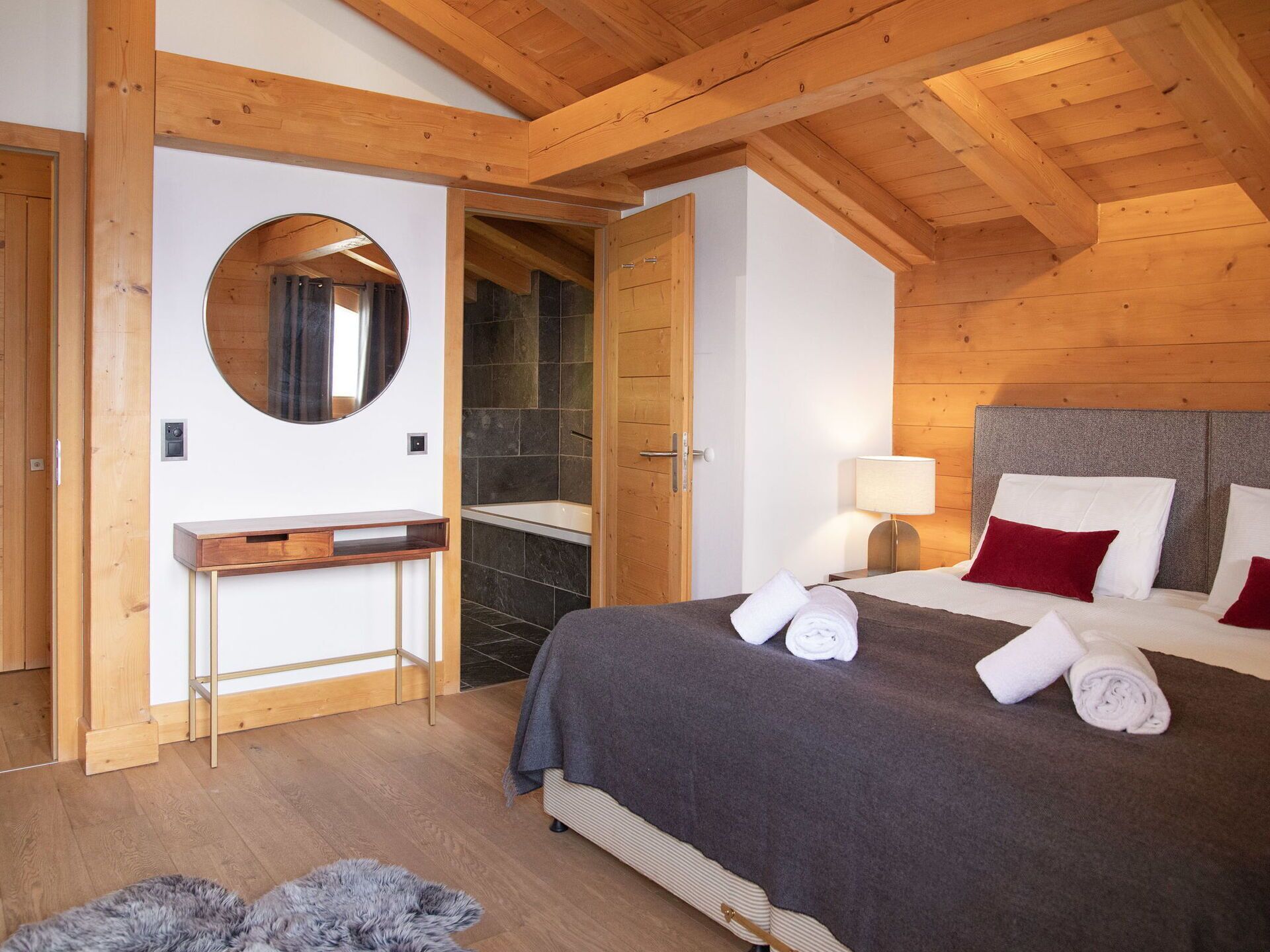 A bedroom with a wooden ceiling, a bed with white and red pillows, a bedside table, a mirror, and a bathroom with a bathtub and tiled walls.
