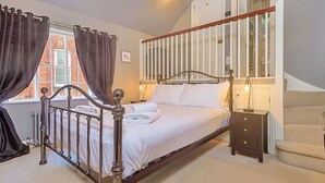 A neatly made bed with white linens, a wooden headboard, and a nightstand with a lamp. A staircase with a wooden railing is visible in the background.