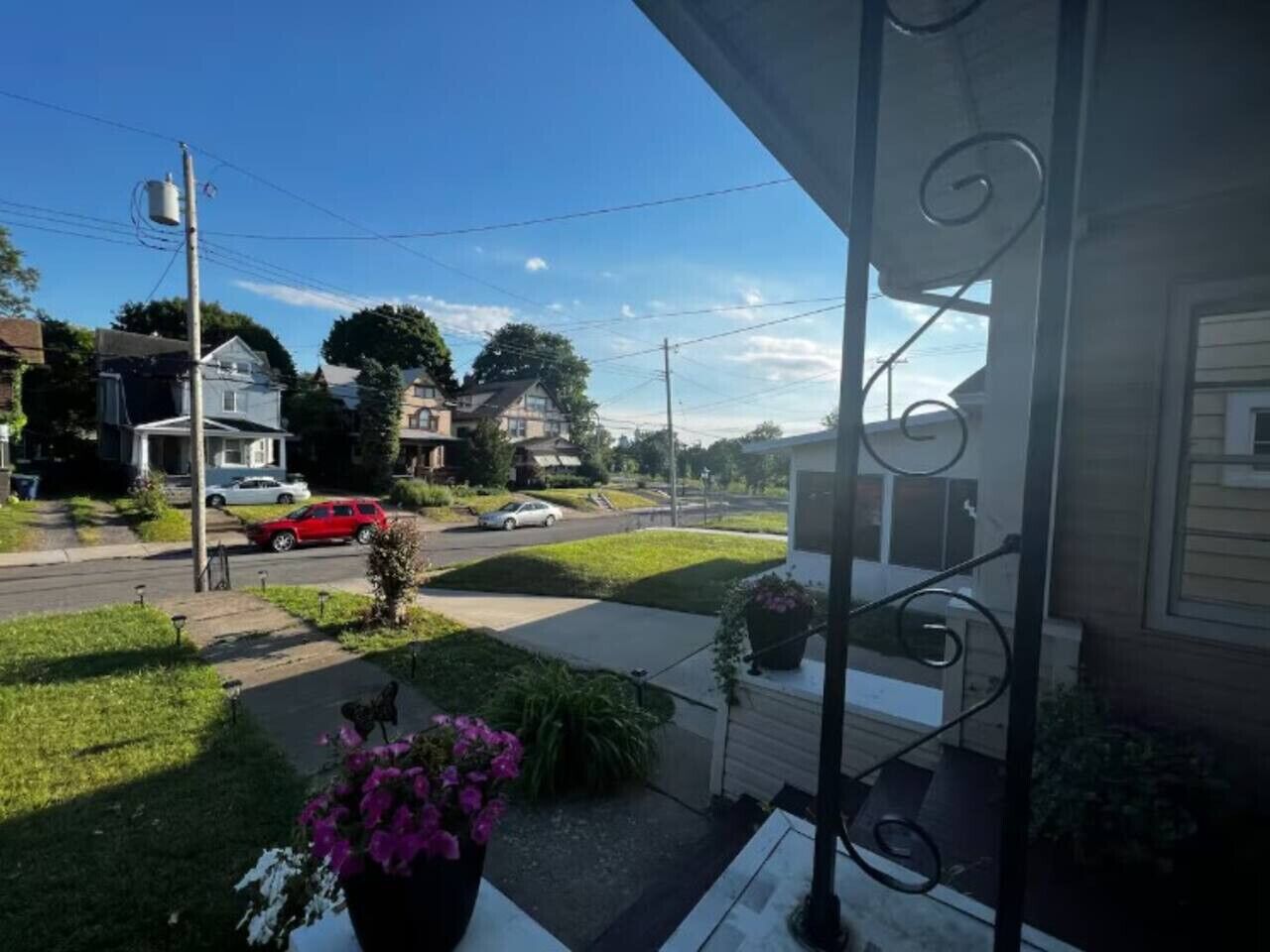 A residential street with houses, a red car parked on the side, and a balcony with a potted plant.