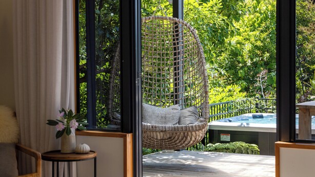 A wicker hanging chair with cushions, a wooden deck, a small table with a vase and a decorative item, and a view of a hot tub through large windows.