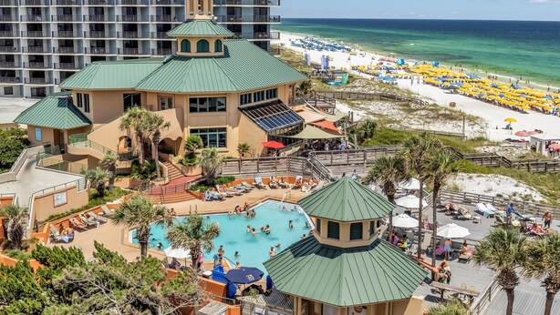Un resort con piscina, zona de playa con sombrillas y un edificio grande al fondo.