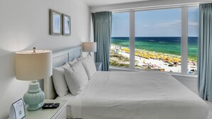 Habitación de hotel con cama, mesita de noche con lámpara, cuadro enmarco y vista a una playa con sombrillas.