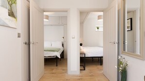 A hallway with two bedrooms, each with a bed and a bedside table. There is a mirror on the right wall and a plant on the left.