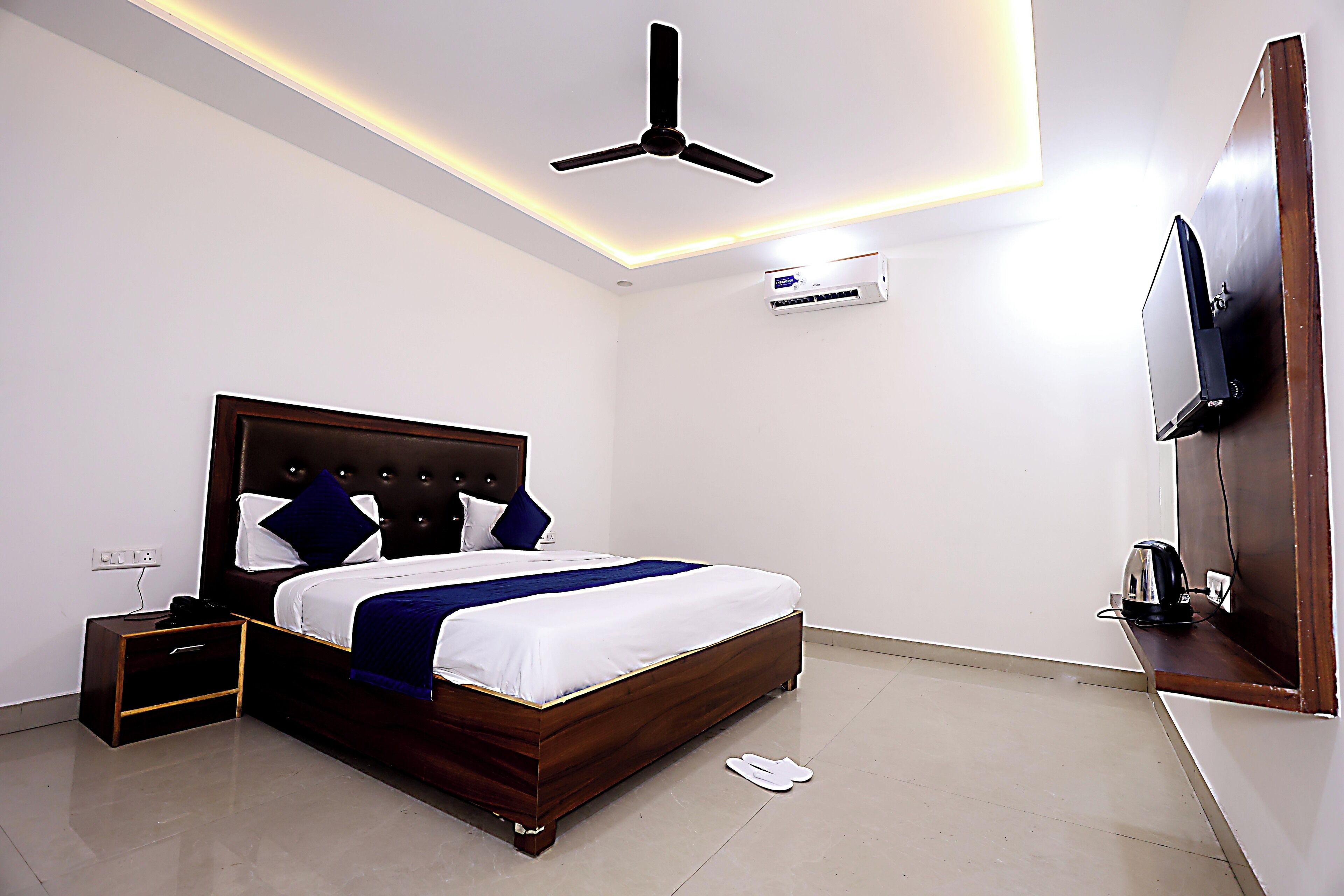 A modern hotel room with a wooden bed, a ceiling fan, an air conditioning unit, a flat-screen TV mounted on the wall, and a small bedside table with a kettle.