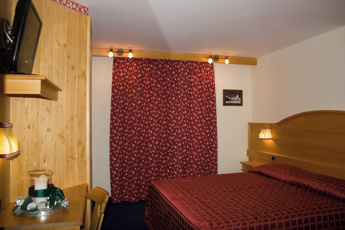 Ein Schlafzimmer mit einem hölzernen Kopfteil, einem Bett mit einem rot gemusterten Bettbezug, einem Nachttisch aus Holz mit einer Lampe, einem an der Wand montierten Regal mit einem Fernseher und einem Vorhang mit einem floralen Muster.