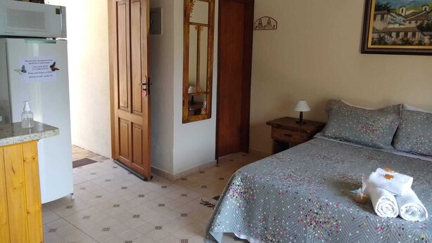 A bedroom with a bed, a nightstand, a mirror, a door, and a kitchenette area with a microwave. - Chalé Topázio (Monte Verde)