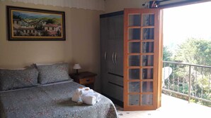 A bedroom with a bed, a nightstand, a dresser, and a balcony with a view of trees. - Chalé Topázio (Monte Verde)
