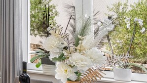 A window with a view of a building and trees, a bottle of wine on a table, and a potted plant with white flowers.
