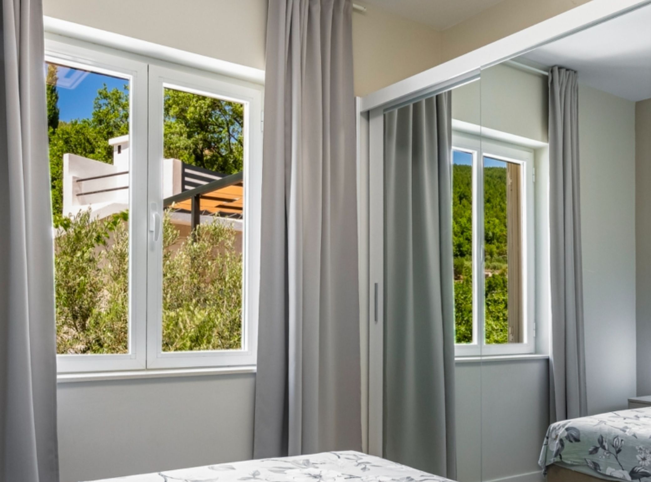 A bedroom with two beds, large windows with curtains, and a view of greenery and buildings outside.