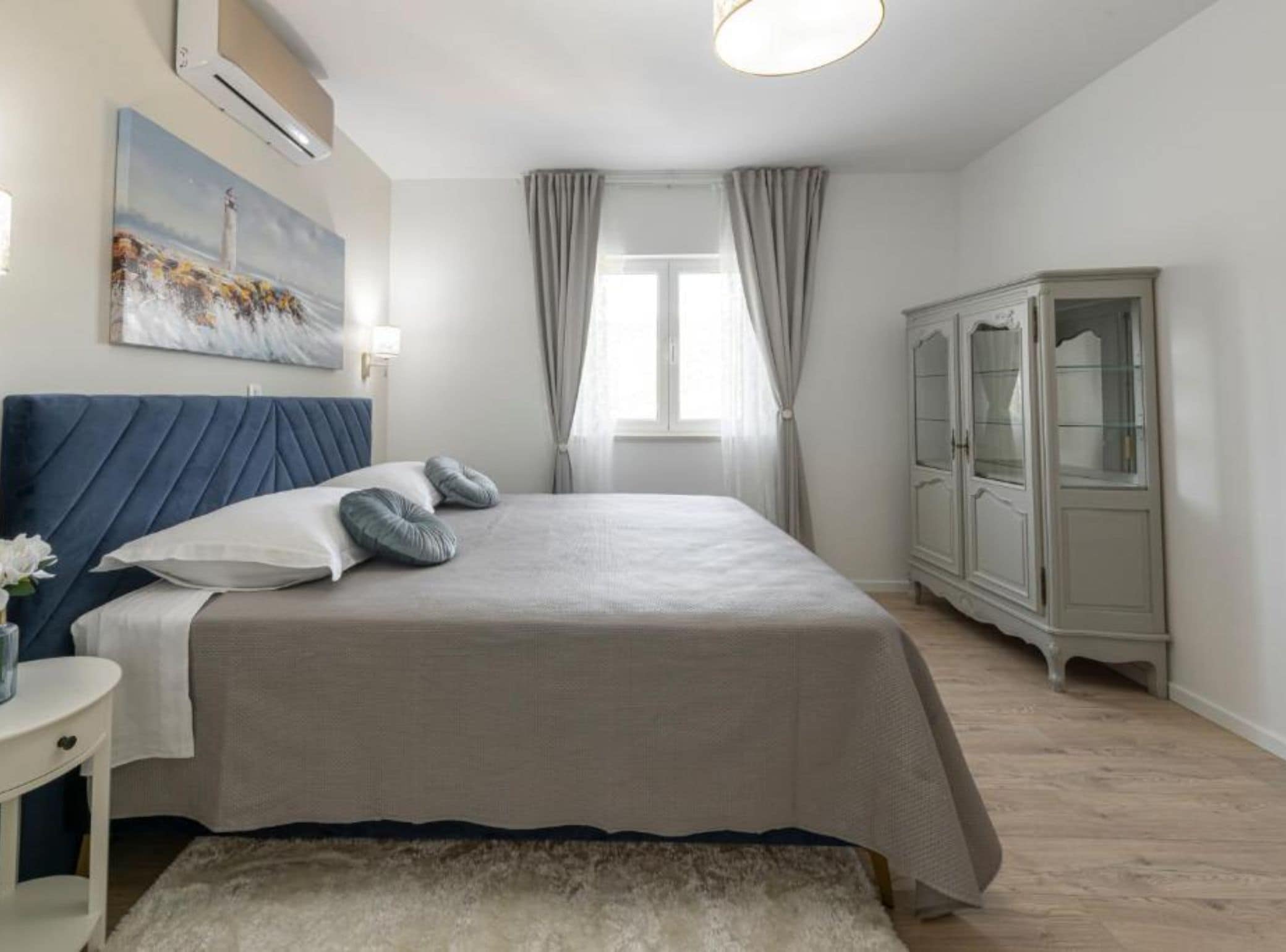 Un dormitorio con cama, mesita de noche, armario, un cuadro y ventana con cortinas.