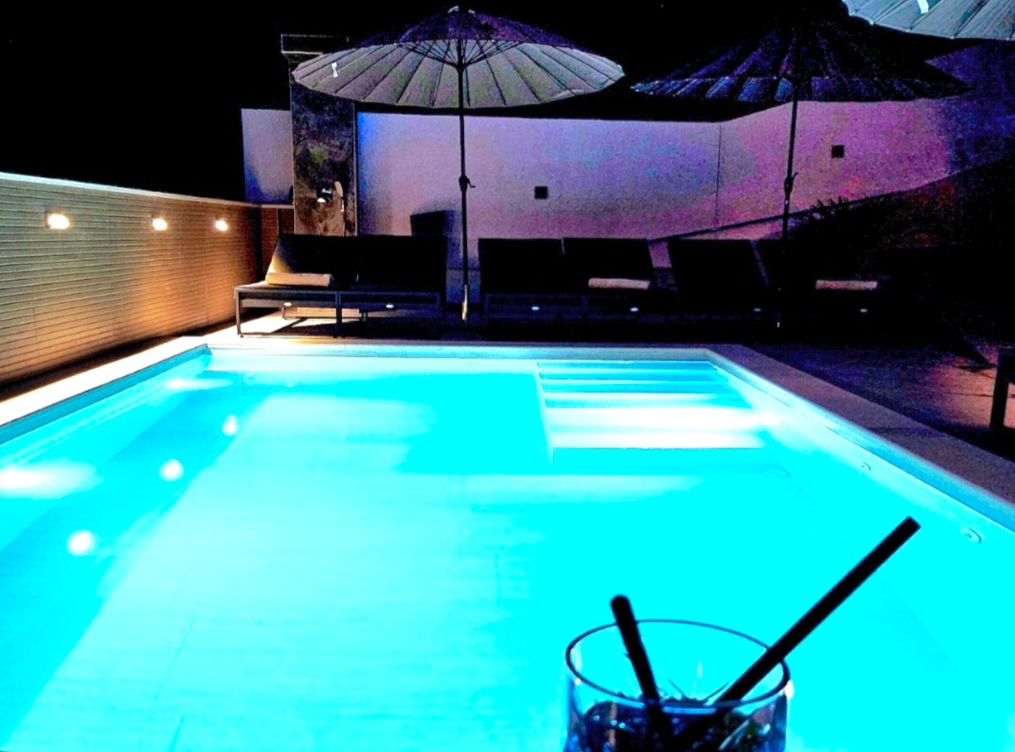 A swimming pool with a glass of water and straws, surrounded by lounge chairs and umbrellas.