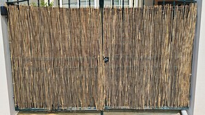 Une clôture en bambou avec un portail en métal devant un bâtiment doté d’un toit en tuiles et d’une fenêtre avec des volets.