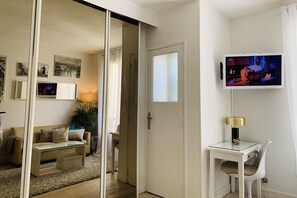 Uma sala de estar moderna com um sofá, uma mesa de centro de vidro, uma TV fixada na parede e uma porta que dá para outro cômodo.