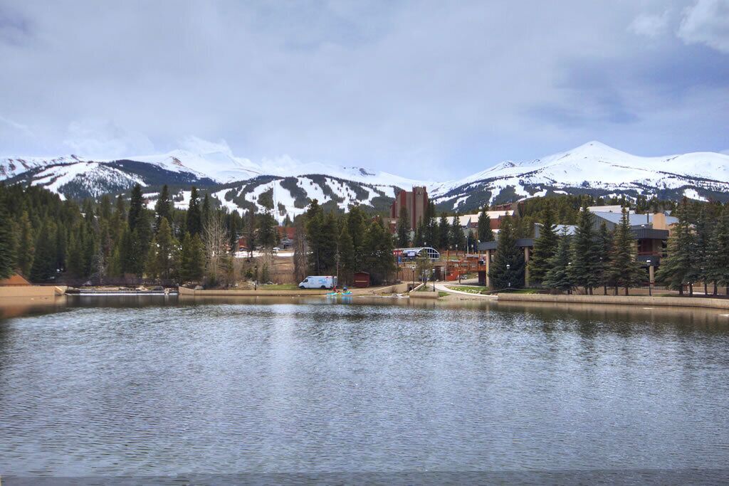 A lake with mountains in the background, surrounded by buildings and trees.
