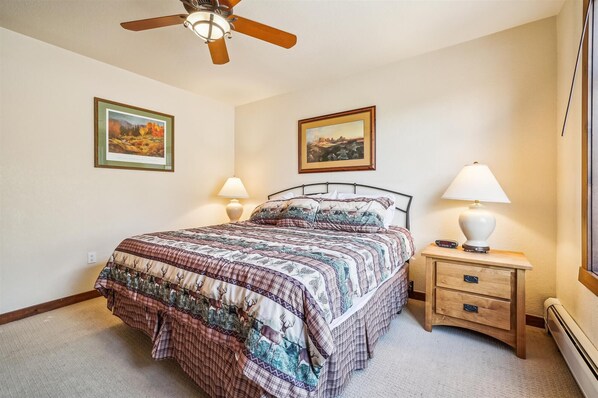 A bedroom with a bed, two bedside tables, a ceiling fan, and two wall-mounted paintings.