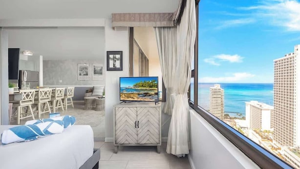 A hotel room with a bed, a TV, a dining area, and a view of the ocean. - Ocean-view condo w/ lanai & sweeping views (Honolulu)