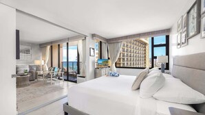 A modern hotel room with a large bed, a seating area with a table and chairs, and a view of a cityscape through large windows. - Ocean-view condo w/ lanai & sweeping views (Honolulu)