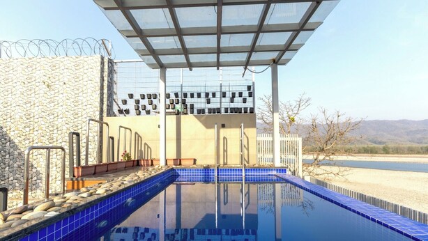 A swimming pool with a stone wall and metal railings, a covered area with a metal roof, and a view of a distant landscape. - Heartland Ganga View Villa Jacuzzi &lift (Rishikesh)