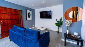 A modern living room with a blue sofa, a TV mounted on the wall, a wooden cabinet, and a small table with a vase and books.