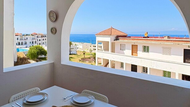 Un coin repas avec une table dressée pour quatre personnes, une vue sur la mer et des bâtiments en arrière-plan.
