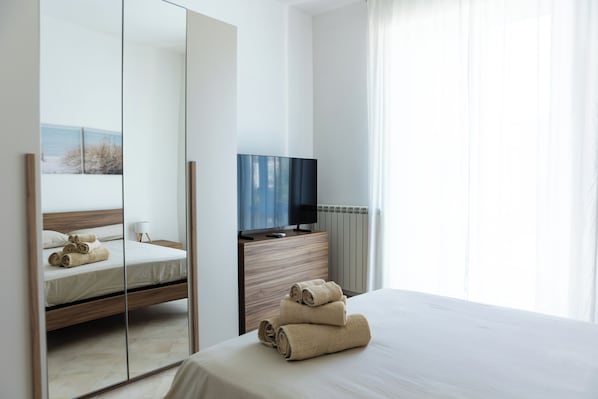 Una camera d'albergo moderna con un letto grande, una televisione su una cassettiera in legno e uno specchio che riflette l'ambiente.