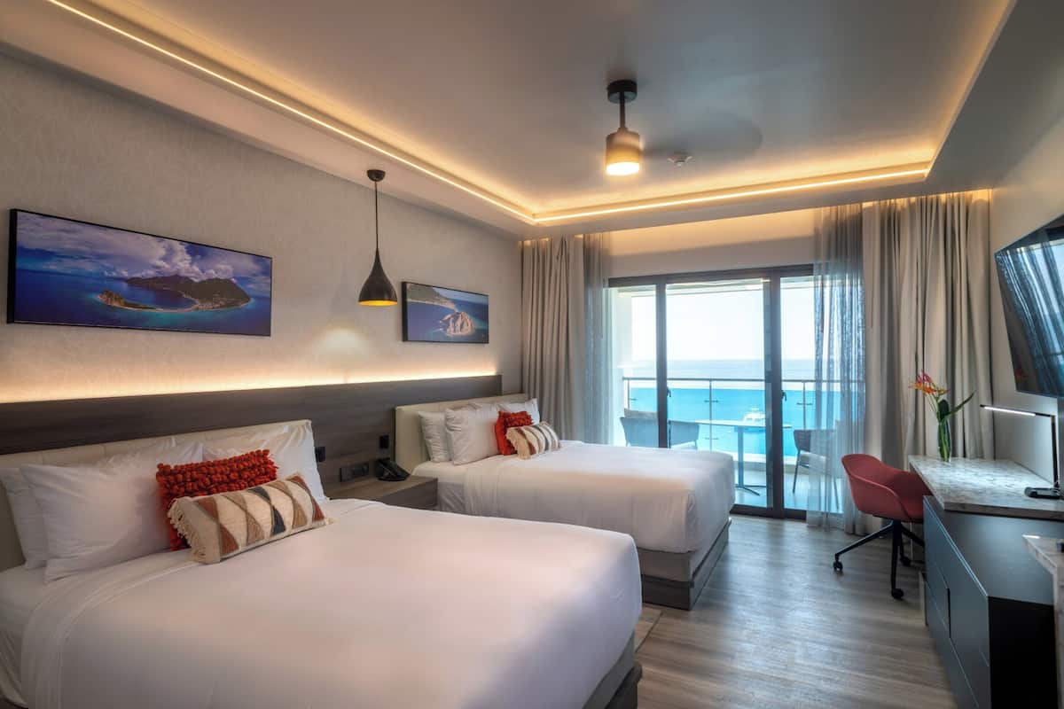 A hotel room with two beds, a desk with a chair, a TV, and a large window with a view of the ocean.