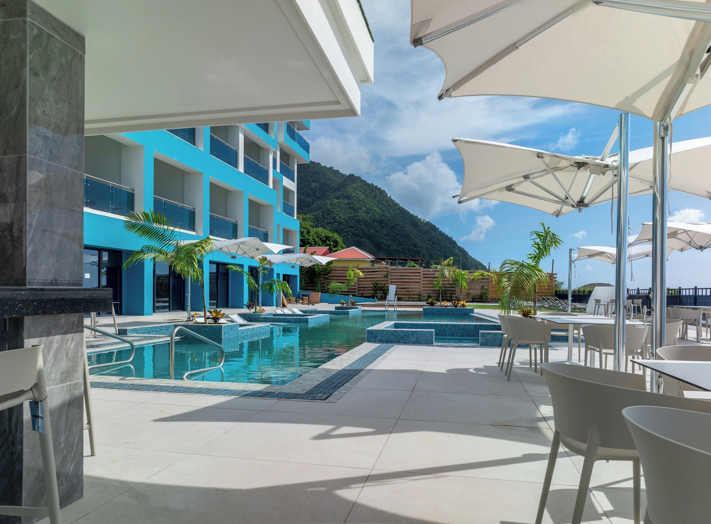 A pool area with multiple swimming pools, lounge chairs, and umbrellas. There are buildings with blue and white exteriors in the background.