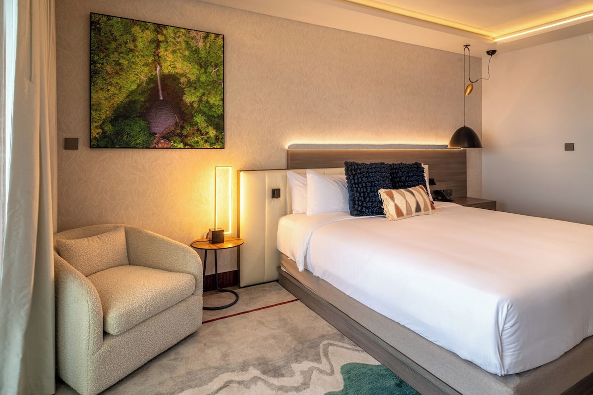 A modern hotel room with a large bed, a small sofa, a bedside table with a lamp, and a wall-mounted artwork.