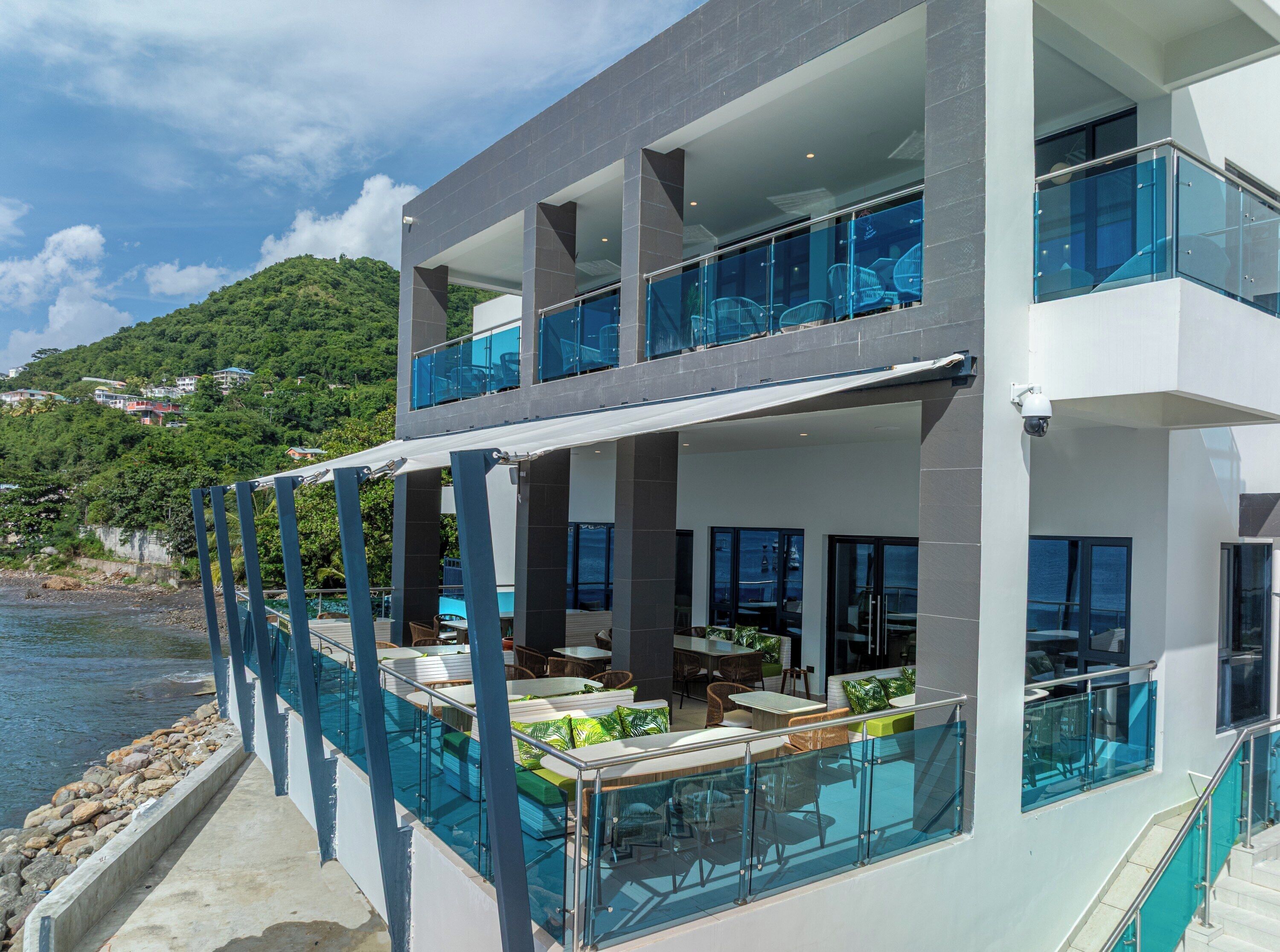 A modern two-story building with a glass balcony overlooking a body of water and a hillside.