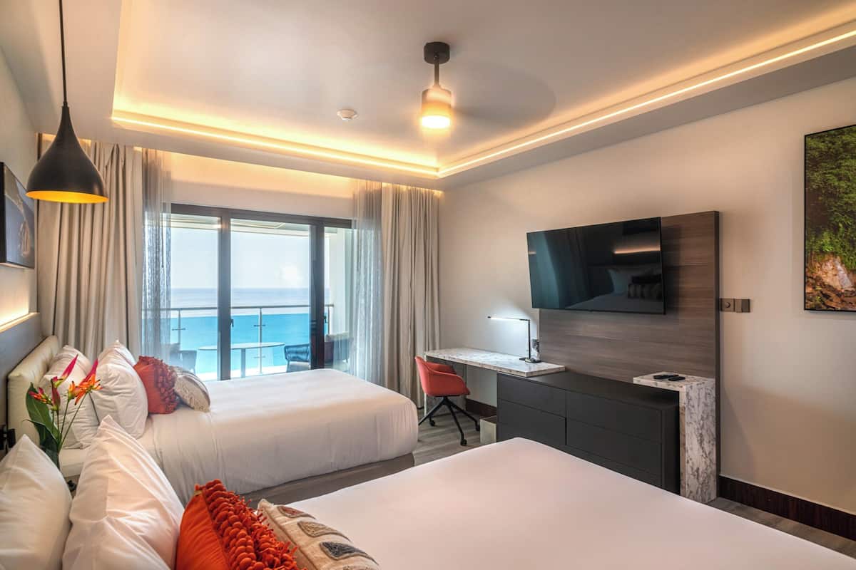 A hotel room with two beds, a desk, a TV, and a view of the ocean.